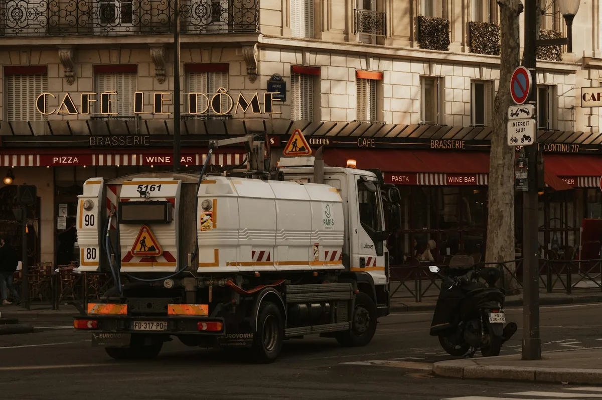 A sanitation truck passes by Cafè Le Dôme on a typical Parisian street, capturing urban life.